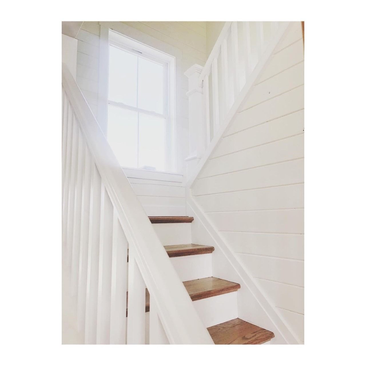 White painted staircase with wooden steps, leading up to a window. Sunlight illuminates the space.