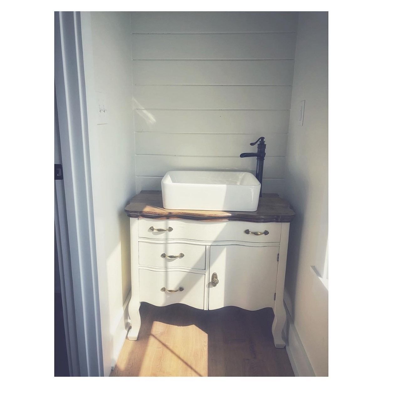 White vanity with sink in a small alcove, antique style with gold hardware, sunlight.