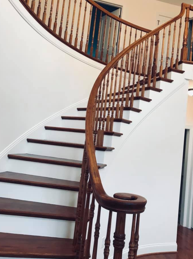 Wooden curved staircase with stained steps and railing against white walls.