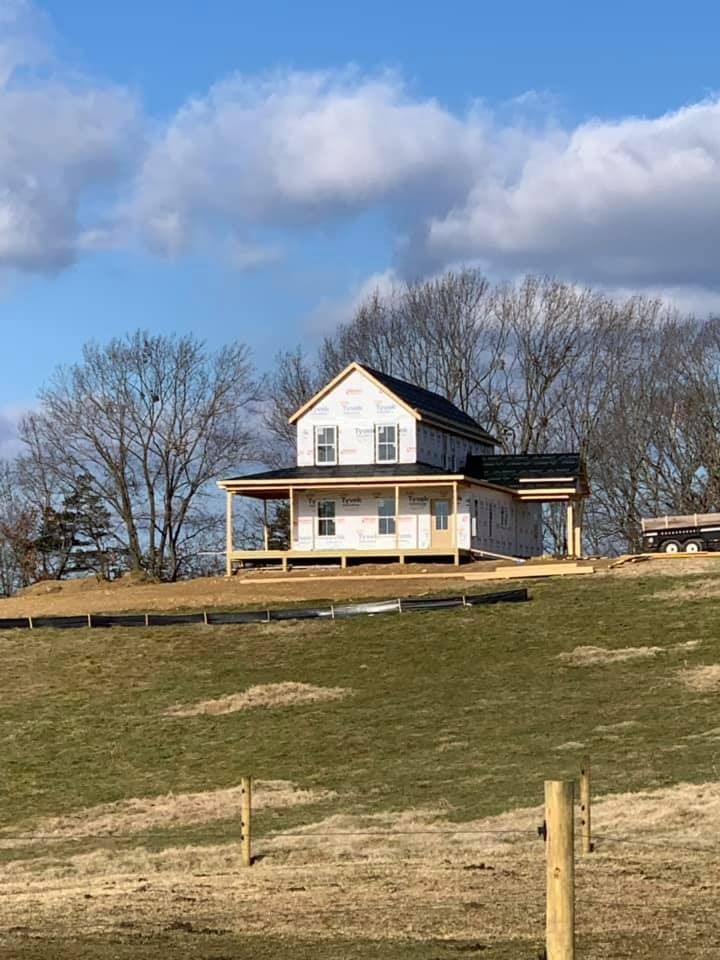 A two-story house under construction on a grassy hill, surrounded by bare trees and a cloudy sky.