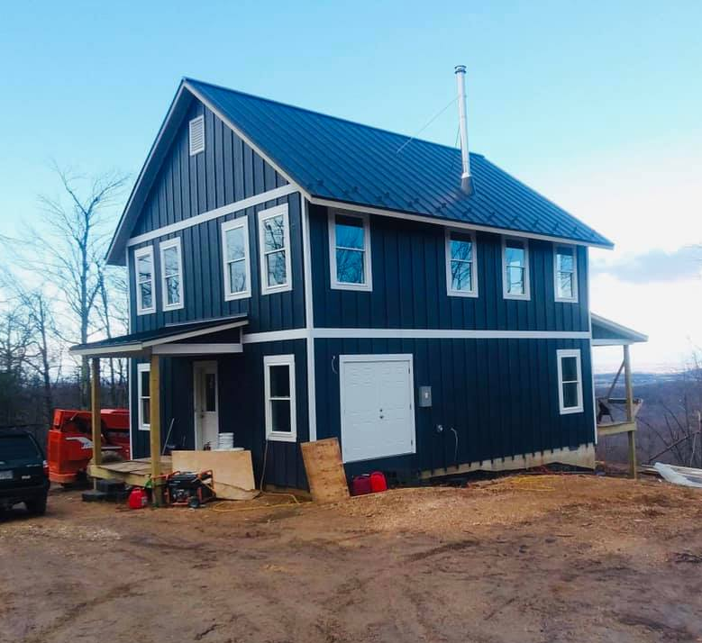 Two-story blue house with white trim, porch, and a black roof. Construction site setting.