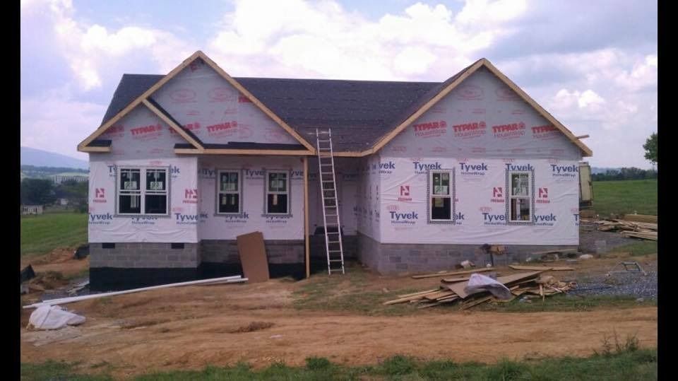House under construction, covered in Tyvek wrap, with ladder leaning against the side; surrounded by construction materials.