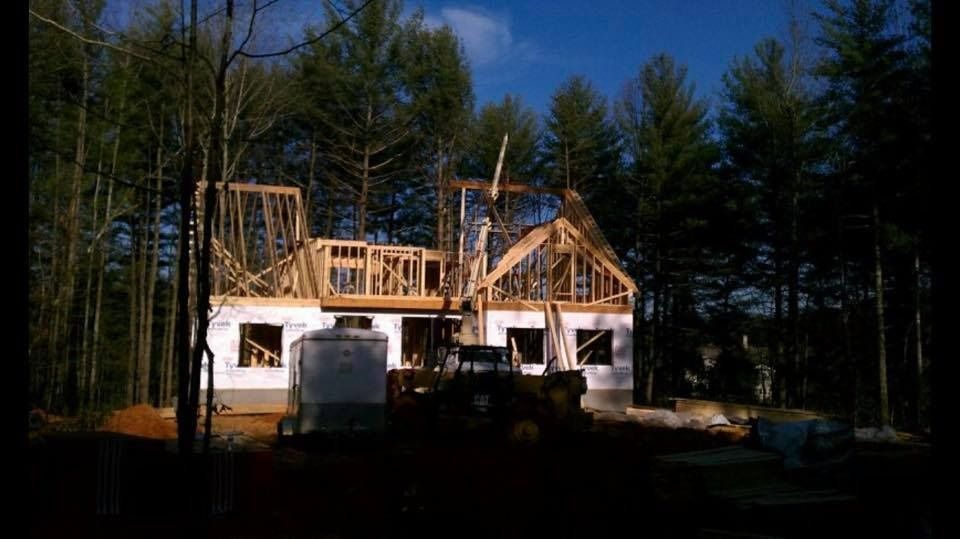 House under construction, wood frame visible. Bulldozer and water tank in front of structure, trees in background.