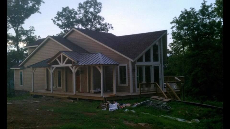 Beige house with a porch and dark roof against a treeline.