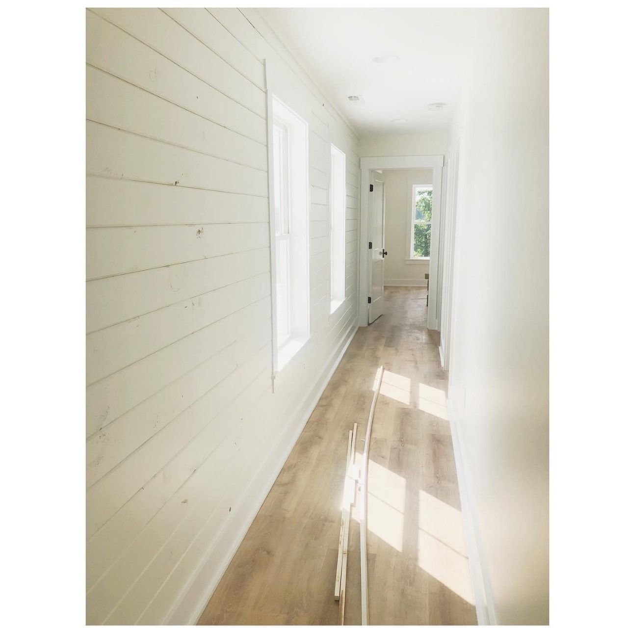 White hallway with shiplap wall, windows, and wood flooring. Sunlight streams in.