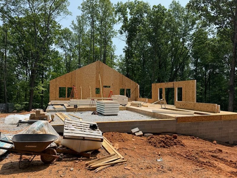 House under construction, wooden frame, surrounded by building materials, set in a wooded area.