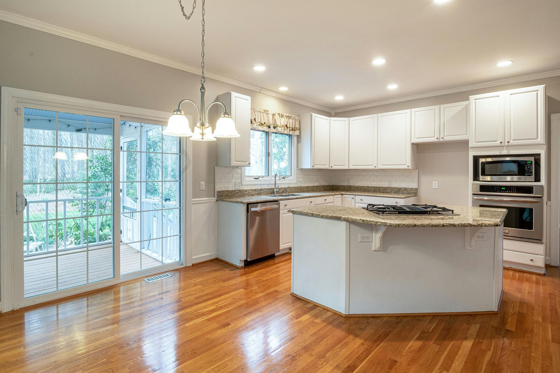 Bright kitchen with white cabinets, island, stainless steel appliances, and wooden floors, leading to a porch.