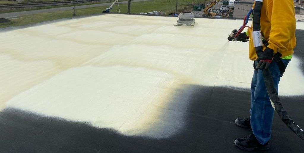 Worker spraying polyurethane foam onto a flat roof surface for insulation and waterproofing.
