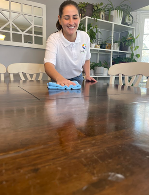 A person in a white polo shirt wipes a dark wooden dining table with a blue cloth in a bright, plant-filled room.