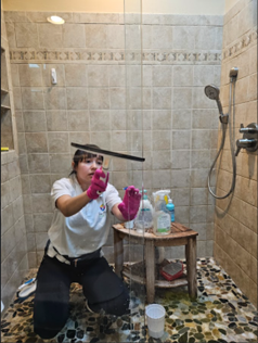 A person wearing pink gloves uses a squeegee to clean a glass shower door inside a tiled shower stall.
