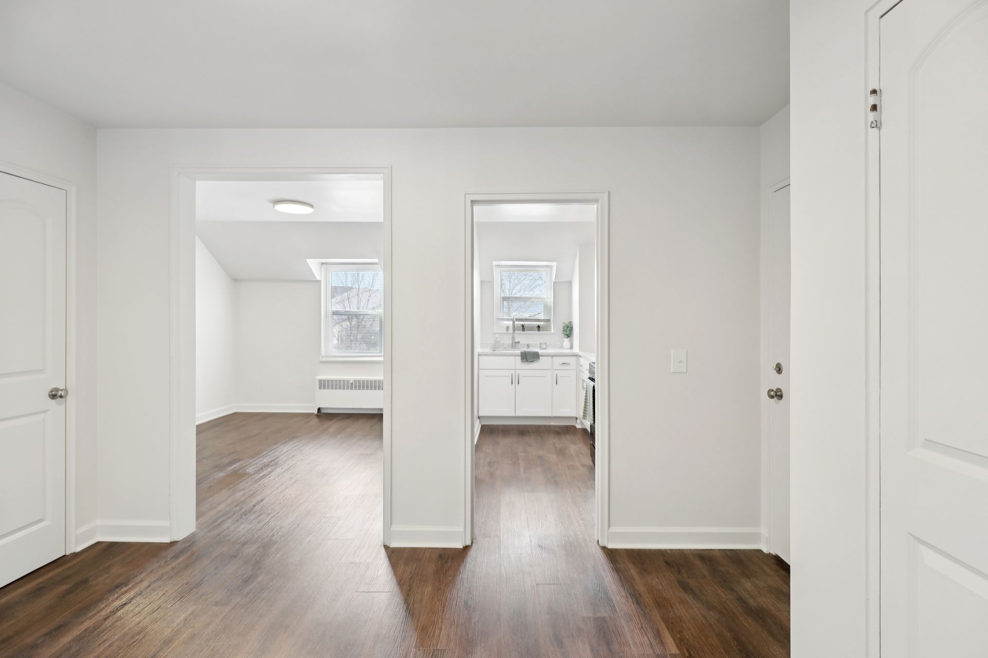 Interior view of a white-walled apartment with three doorways: bedroom, kitchen, and entrance. Dark wood floors.