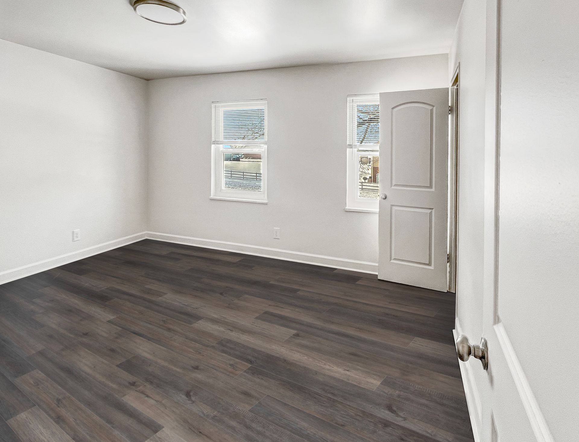 Empty room with dark wood-look flooring, white walls, two windows, and a partially open door.
