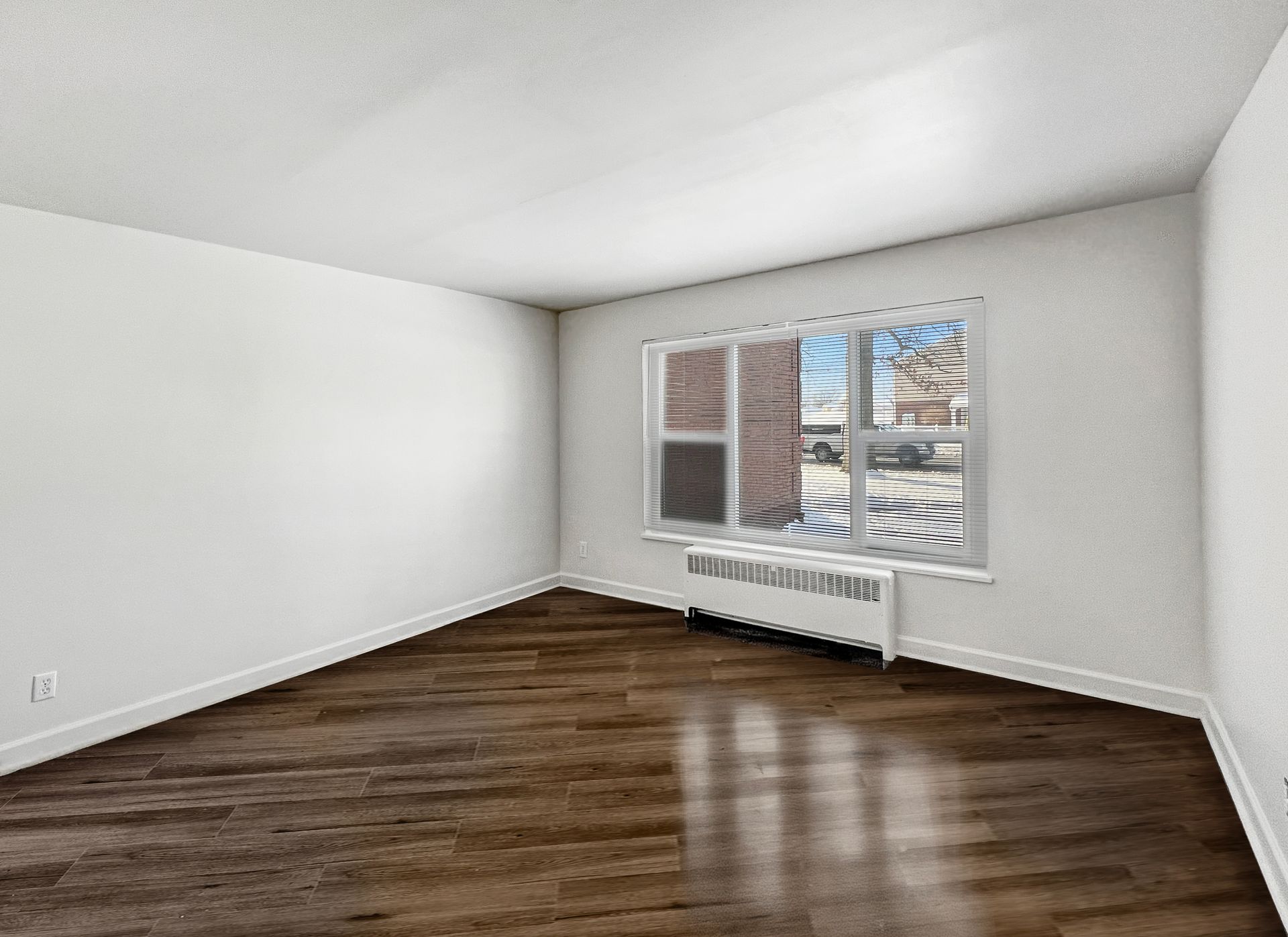 Empty room with dark wood floor, white walls and ceiling, window with blinds.