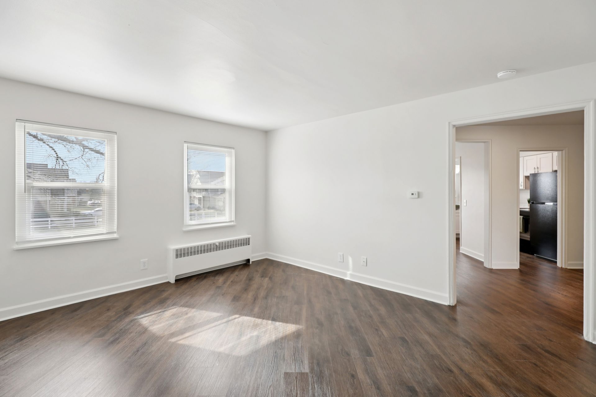 Empty living room with dark wood floor, two windows, and doorway to a kitchen.