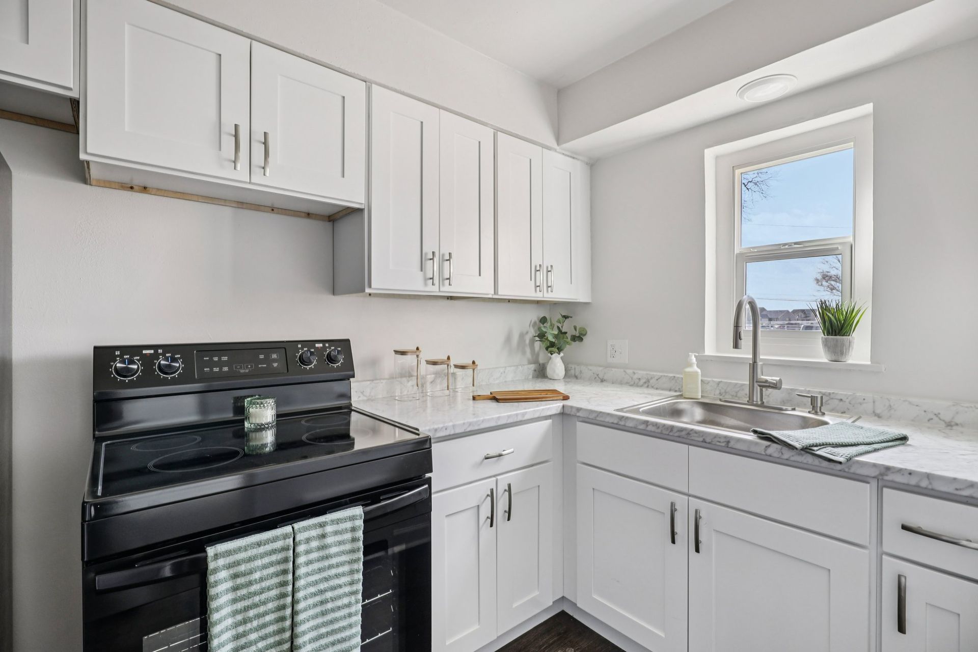 White kitchen with black stove, white cabinets, marble countertops, and a window.