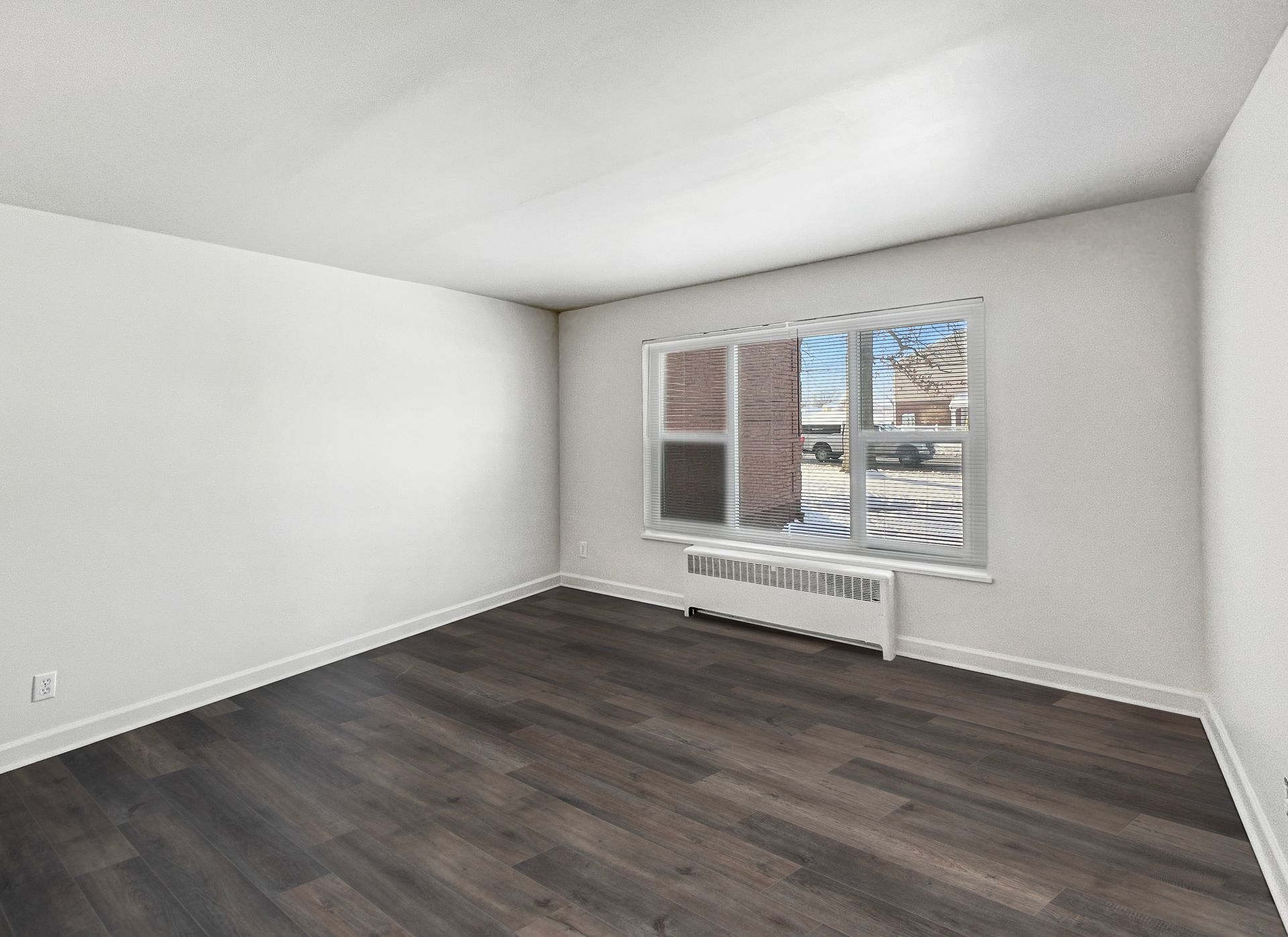 Empty room with dark wood flooring, white walls, window with blinds, and radiator.