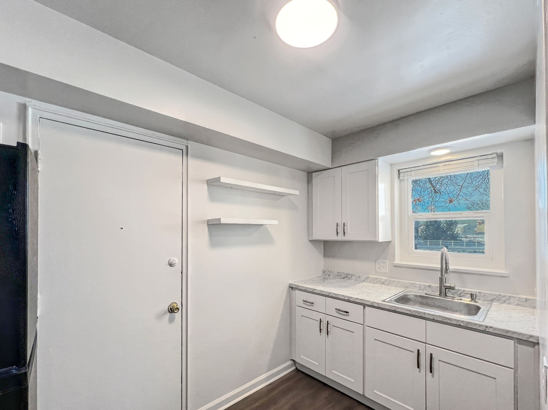 Small white kitchen with cabinets, sink, window, door, and floating shelves.