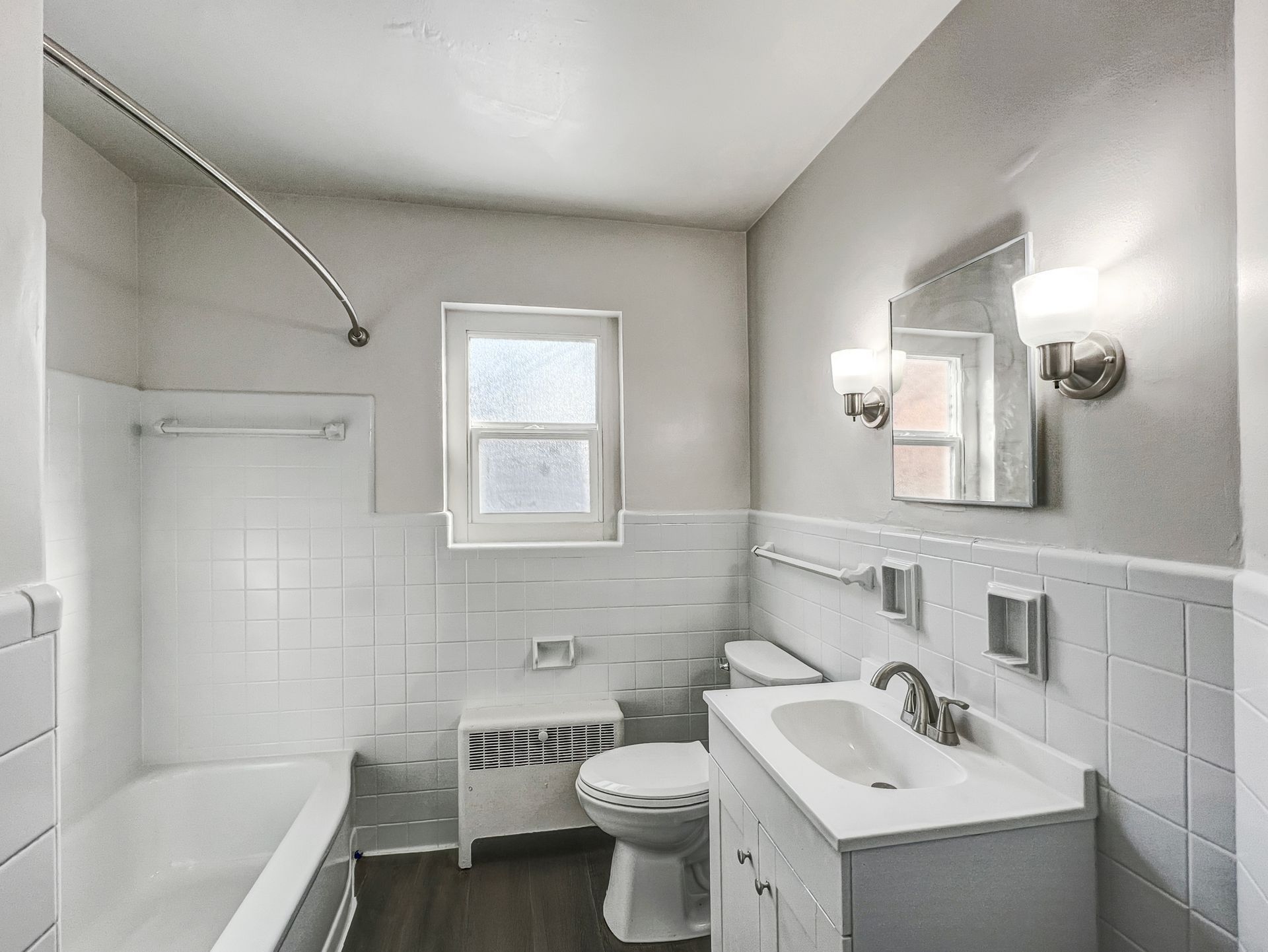 White tiled bathroom with a tub, toilet, and sink. Includes a window and a mirror with sconces.