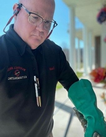 An exterminator wearing glasses, a black uniform, and green gloves, is performing an extermination. An exterminator wearing glasses, a black uniform, and green gloves, is performing an extermination.