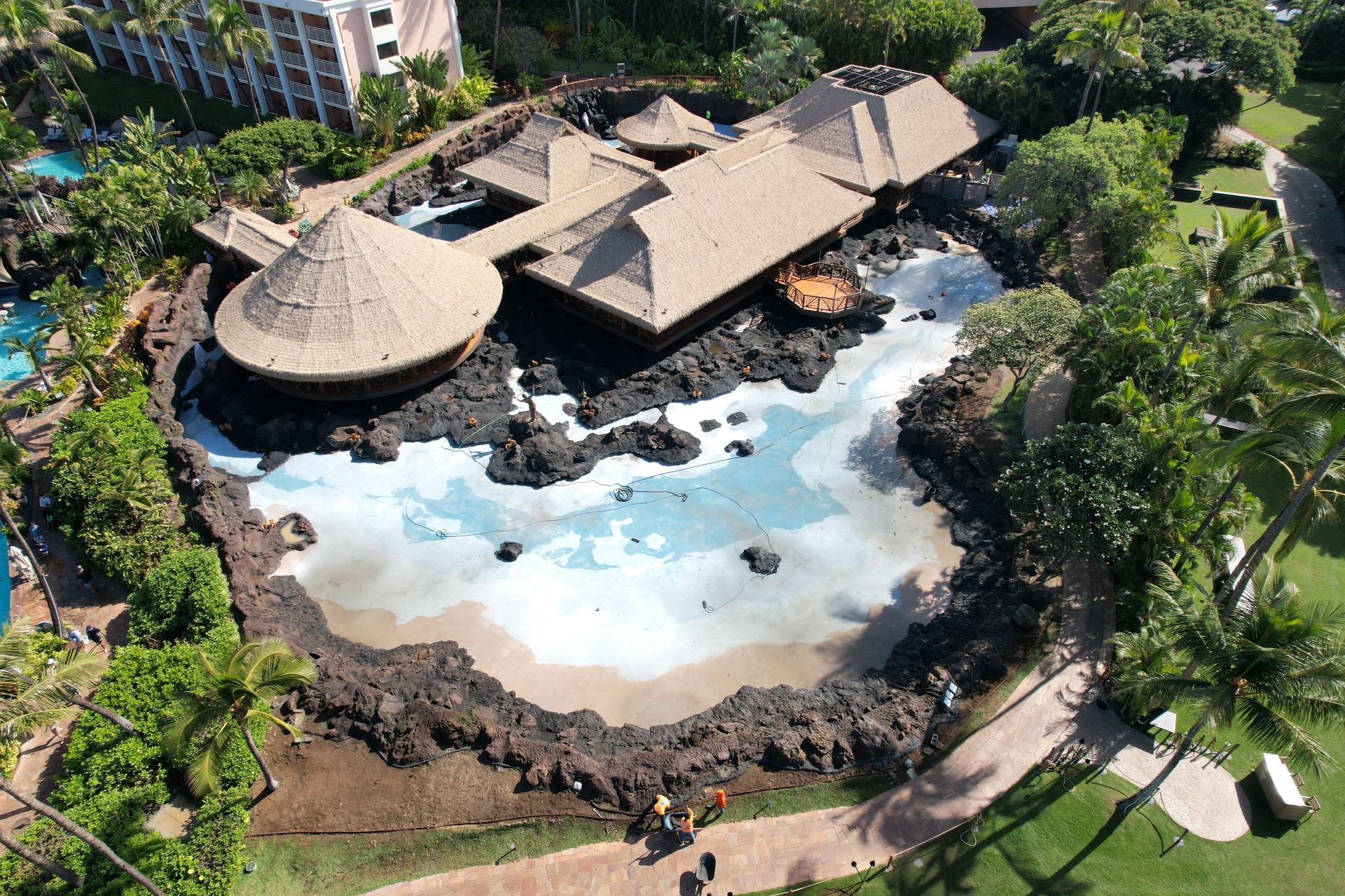 Aerial view of a restaurant with thatched roof, set in a lagoon-like pool surrounded by black rocks and greenery.