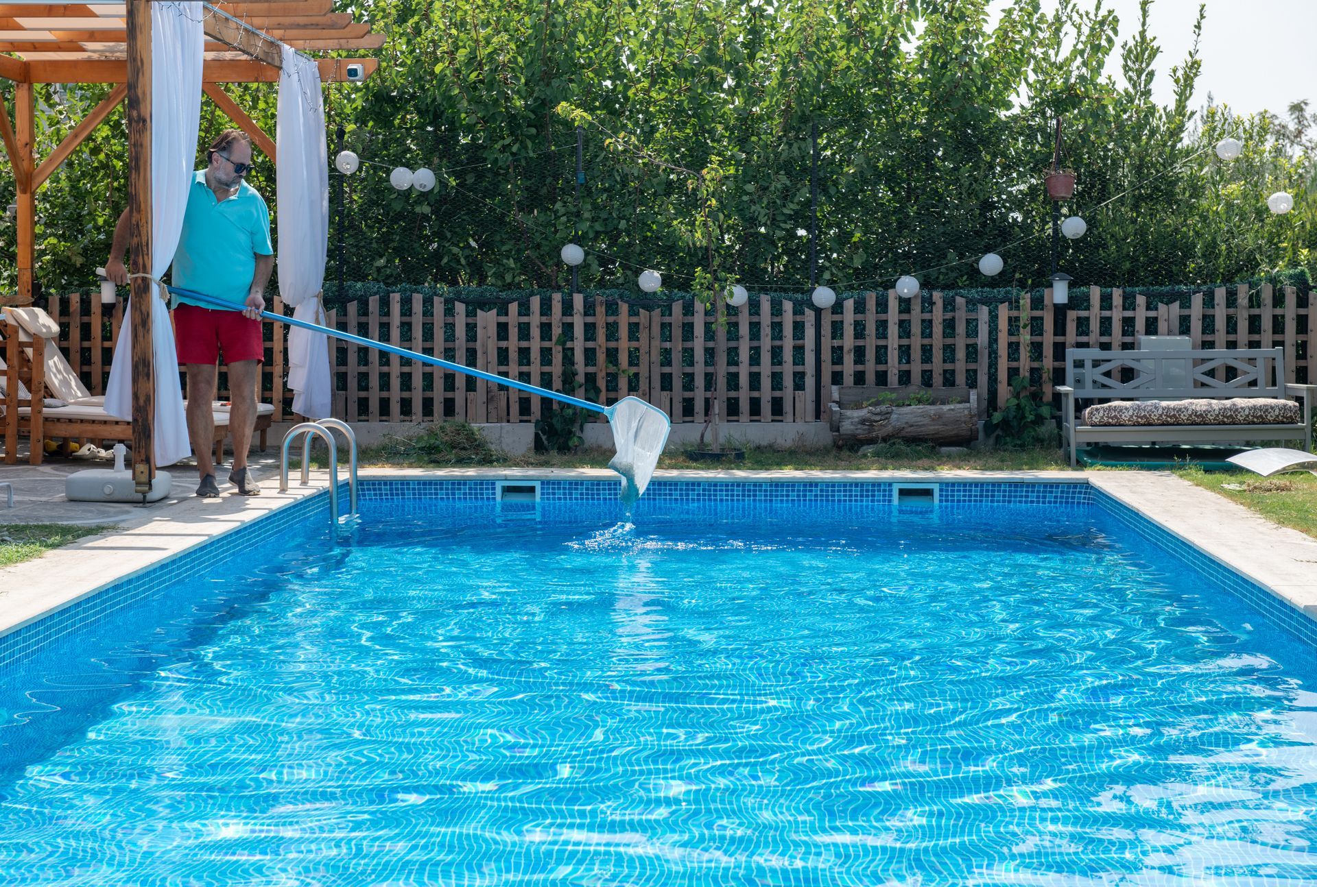 A person in a blue shirt and red shorts cleans a rectangular backyard swimming pool with a long-handled net. A person in a blue shirt and red shorts cleans a rectangular backyard swimming pool with a long-handled net.