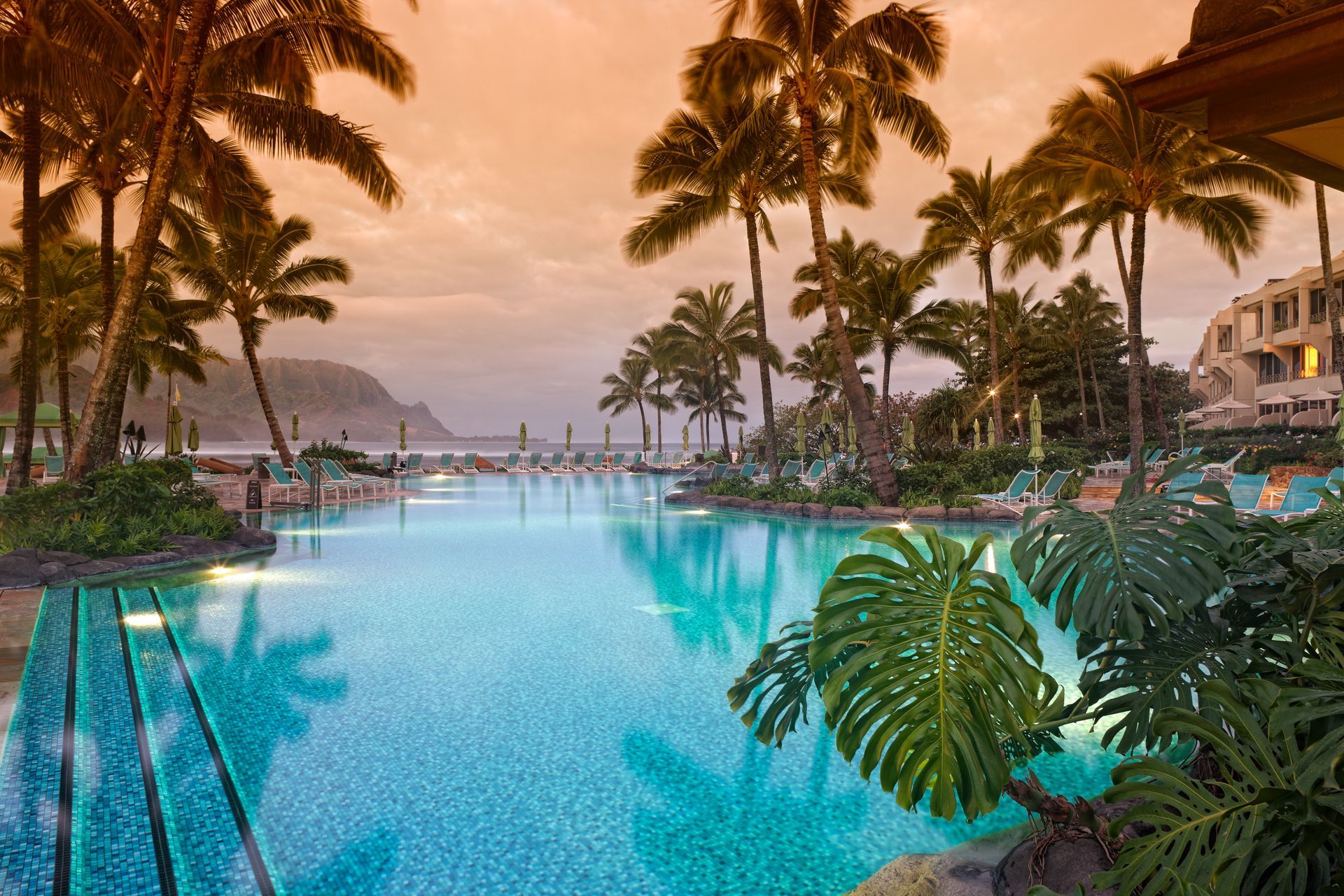 A luxurious resort pool surrounded by palm trees with a mountain view during sunset.