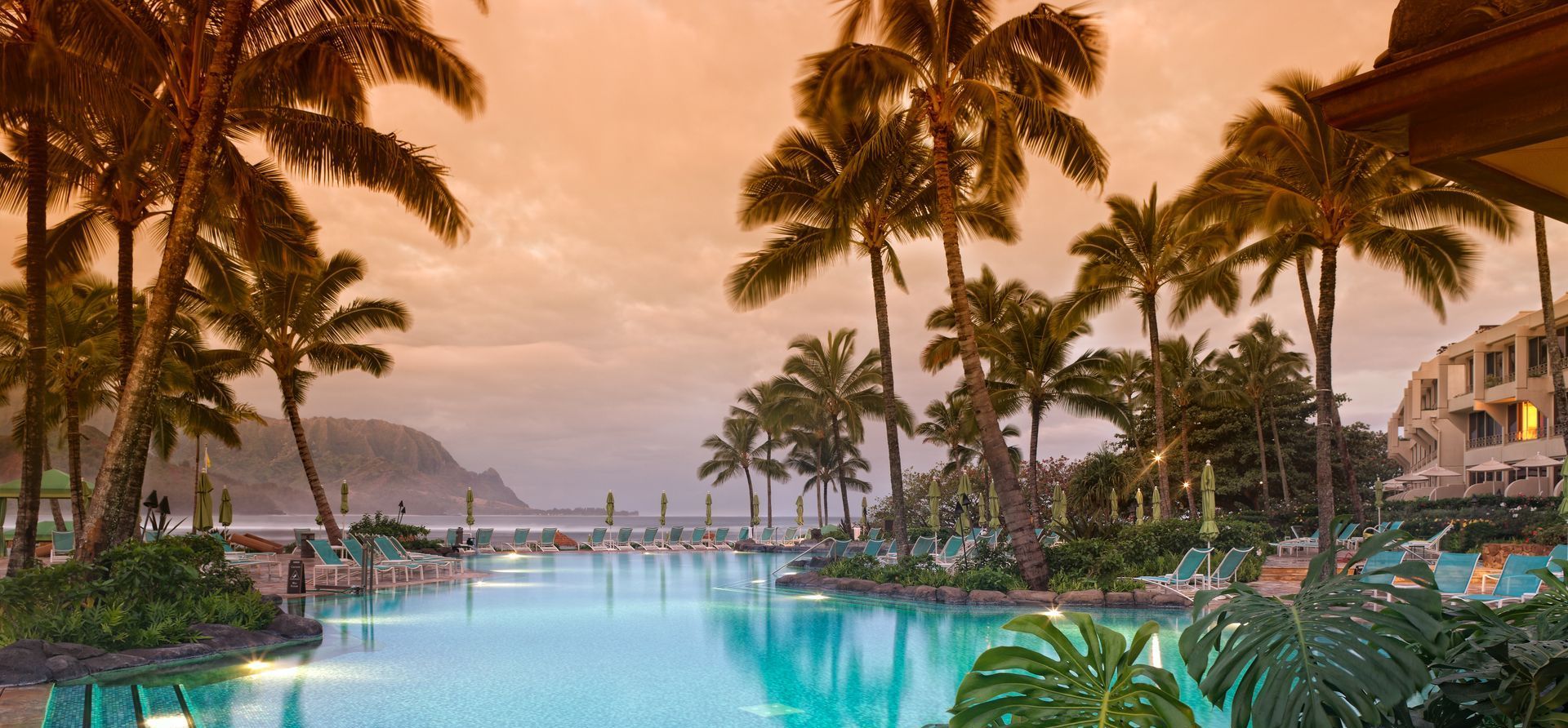 A tropical pool at sunset, with palm trees, mountains, and a resort.