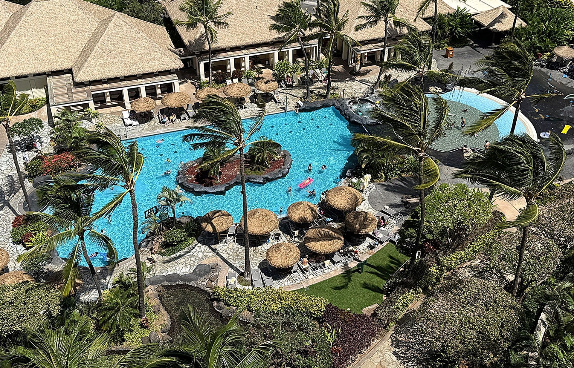 Tropical resort pool with thatched-roof huts, lush greenery, and a small island in the center.