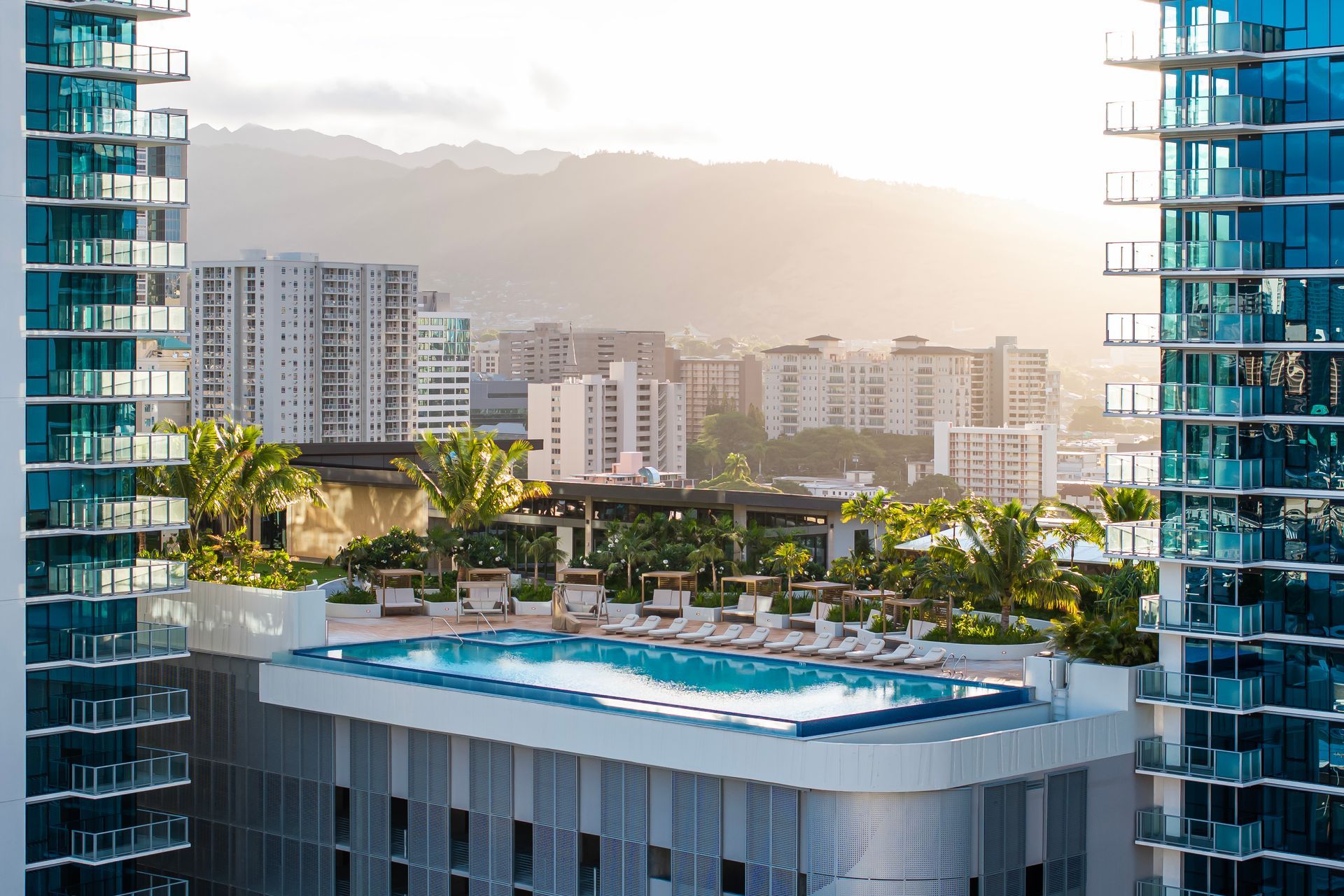 A tropical pool at sunset, with palm trees, mountains, and a resort.