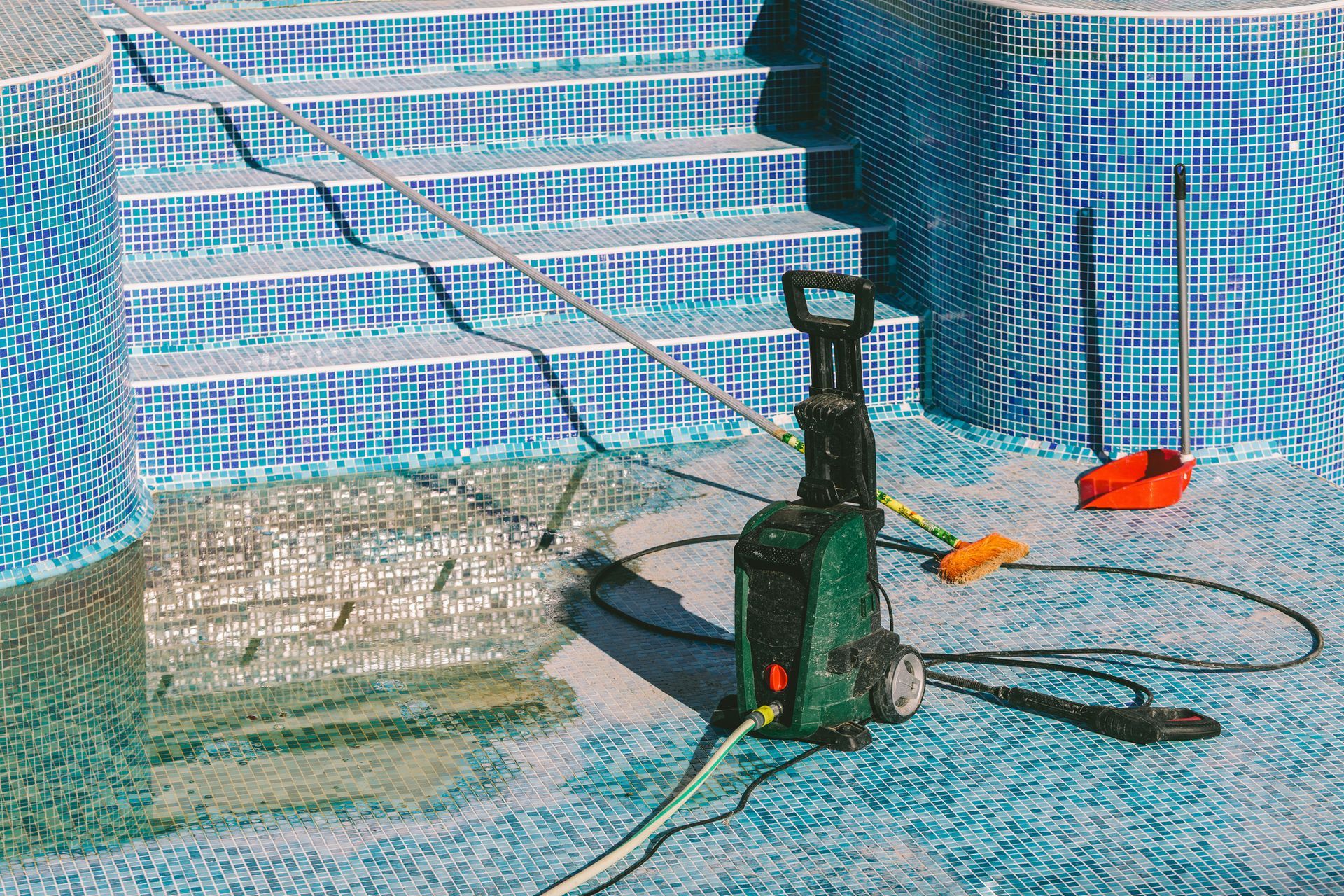 An empty pool with blue tiles being pressure washed, with steps and cleaning tools visible