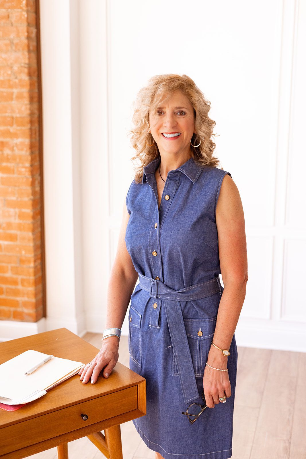 Woman with curly hair smiles, sitting at wooden desk in light-filled room; brick wall, plant visible.