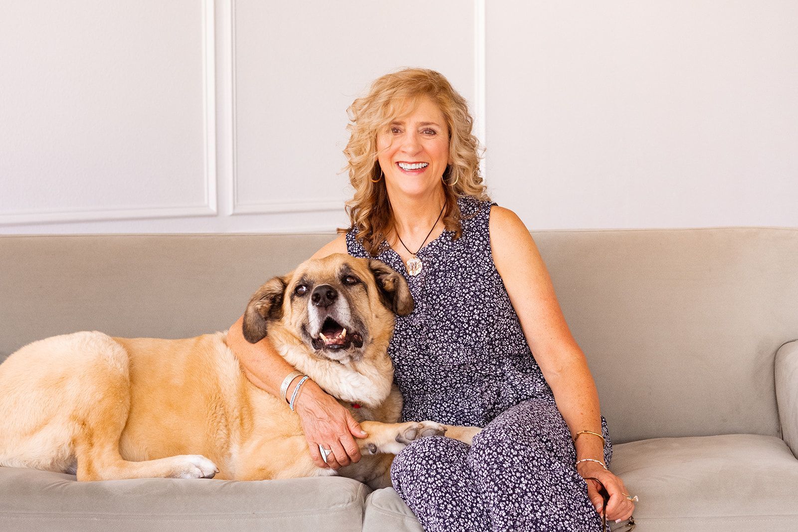 Woman with curly hair and dog sit on a mint green couch. Woman smiles. The dog looks happy. Light wood floor, white wall.