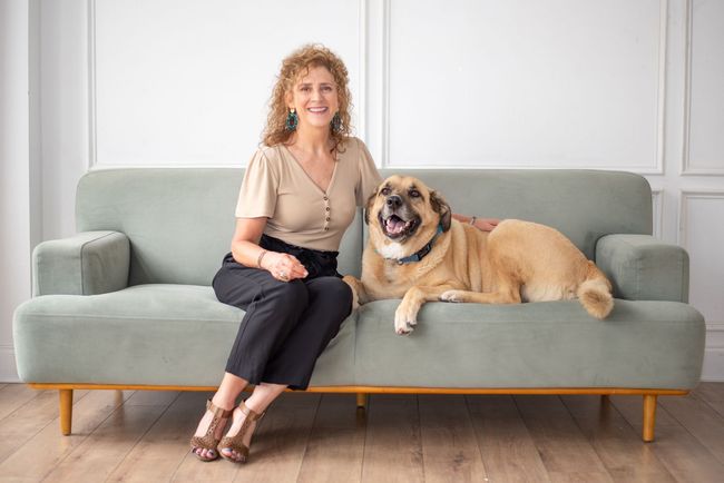 Woman with curly hair and dog sit on a mint green couch. Woman smiles. The dog looks happy. Light wood floor, white wall.