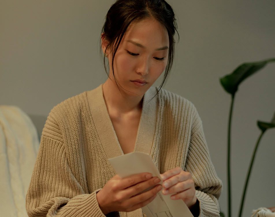 Woman in a tan sweater reading a letter indoors with a plant in the background.