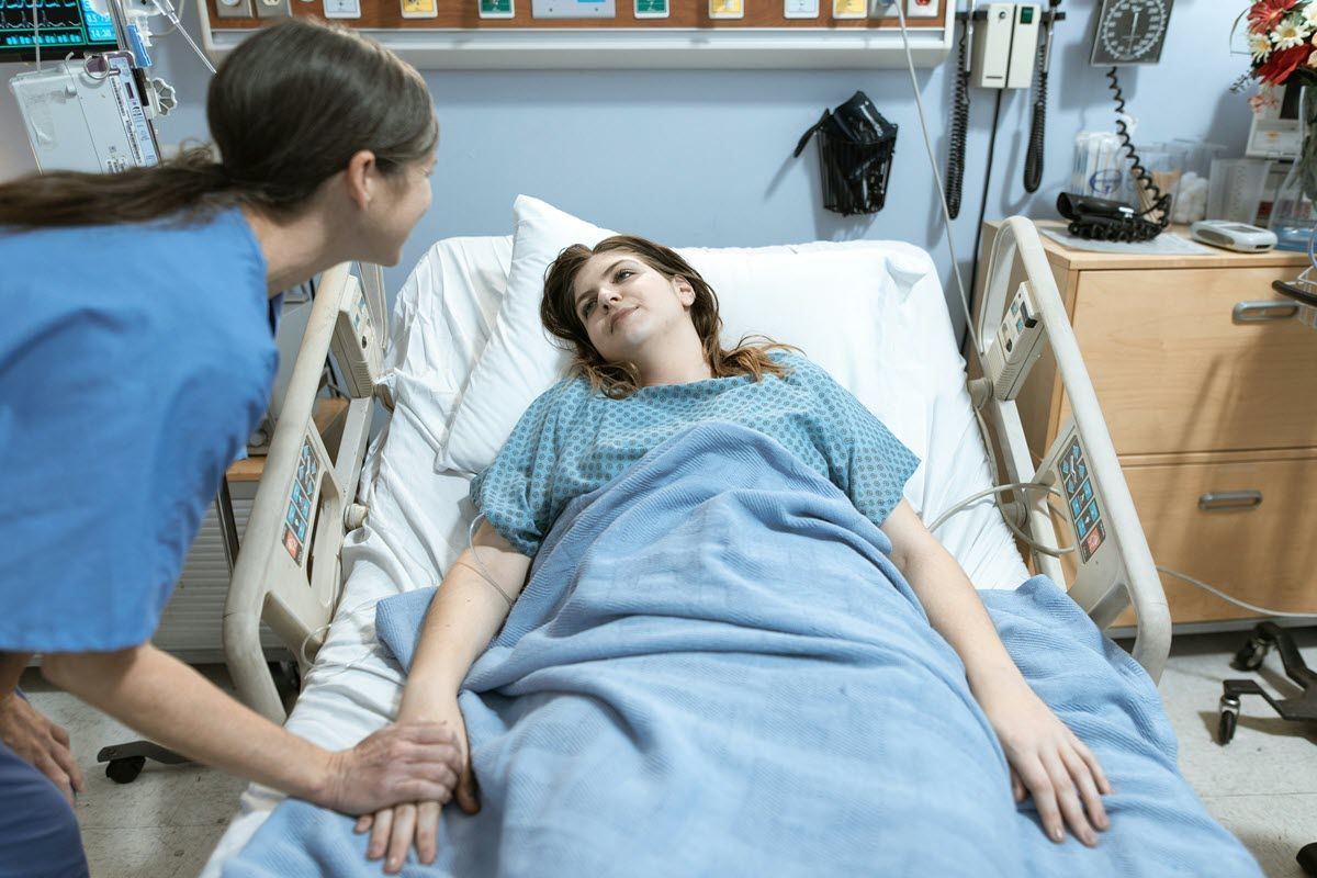 A caregiver in blue scrubs holds the hand of a person resting in a hospital bed with a blue blanket.