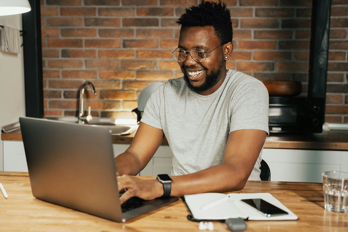 Man smiling while using laptop at kitchen counter. Gray shirt, glasses, watch. Brick wall background.