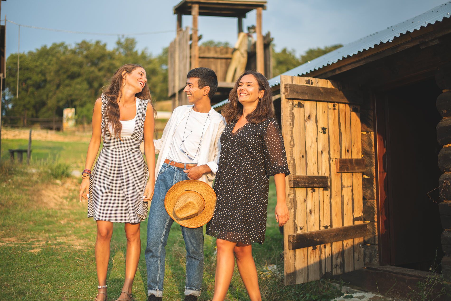 Three people laughing outdoors: a man in denim, two women in dresses; wooden cabin and watchtower in background.