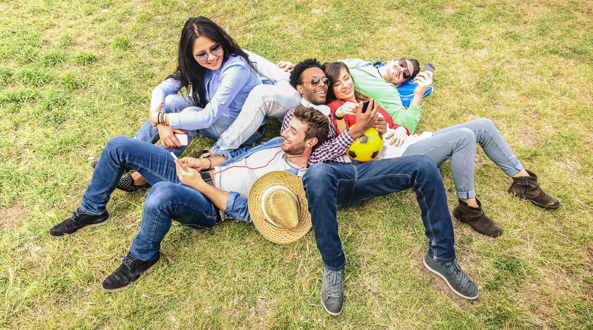 Group of friends lying on grass, smiling and hugging, enjoying a sunny day outdoors.