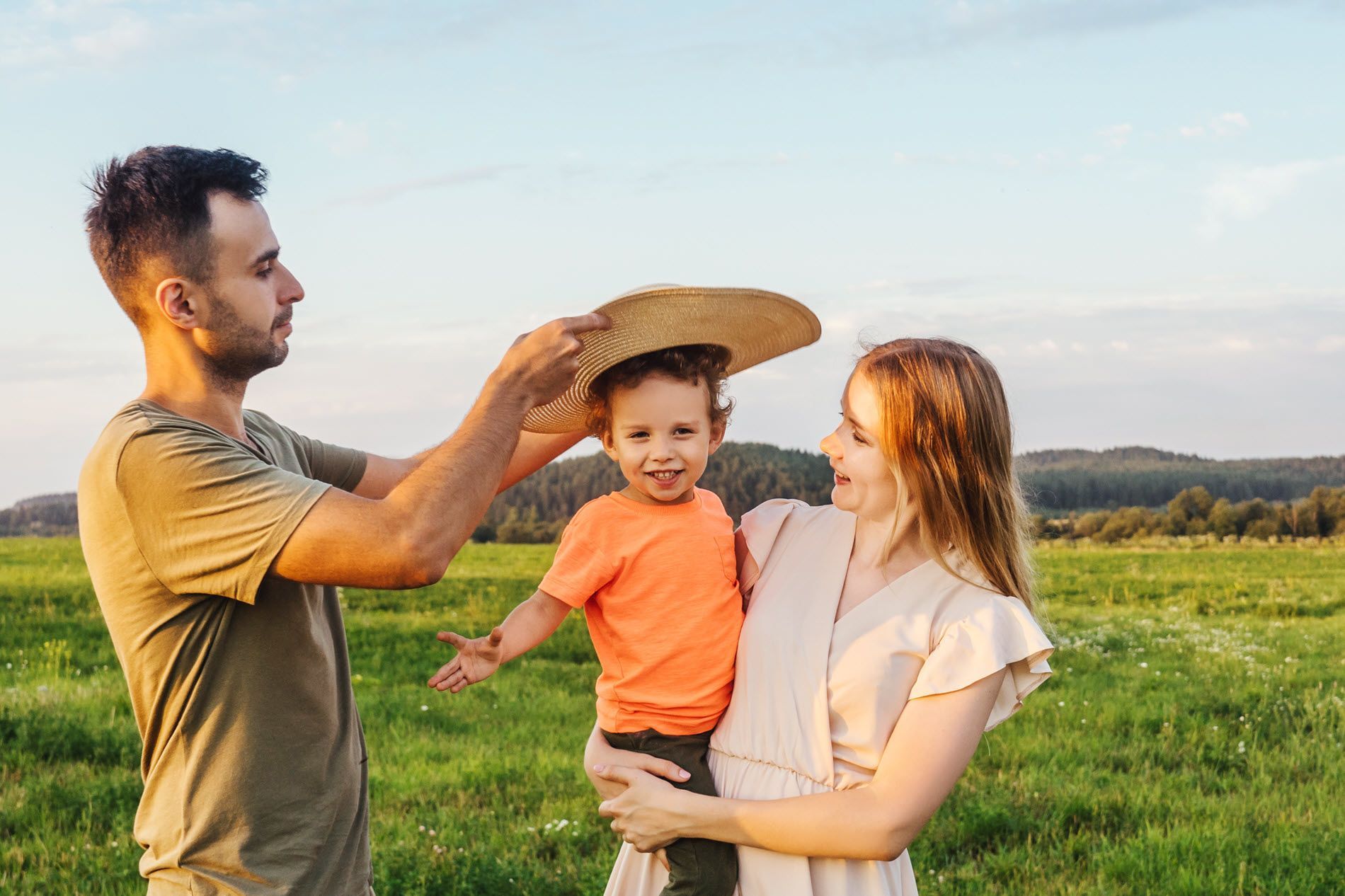 Father putting a straw hat on a smiling toddler held by his mother in a green field.