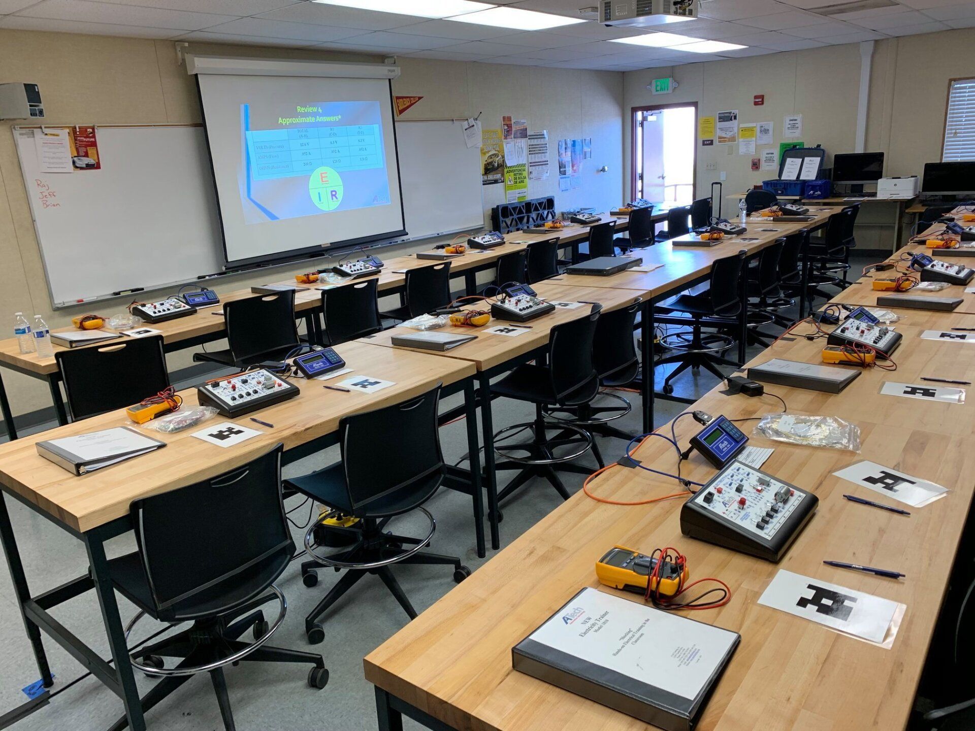 A classroom with a lot of tables and chairs and a projector screen.