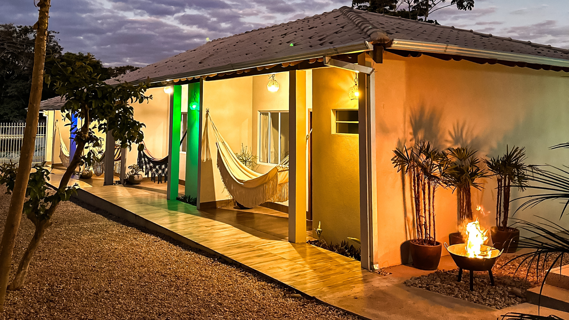 Chalé com varanda coberta ao entardecer, com redes, fogueira e vasos de plantas.
