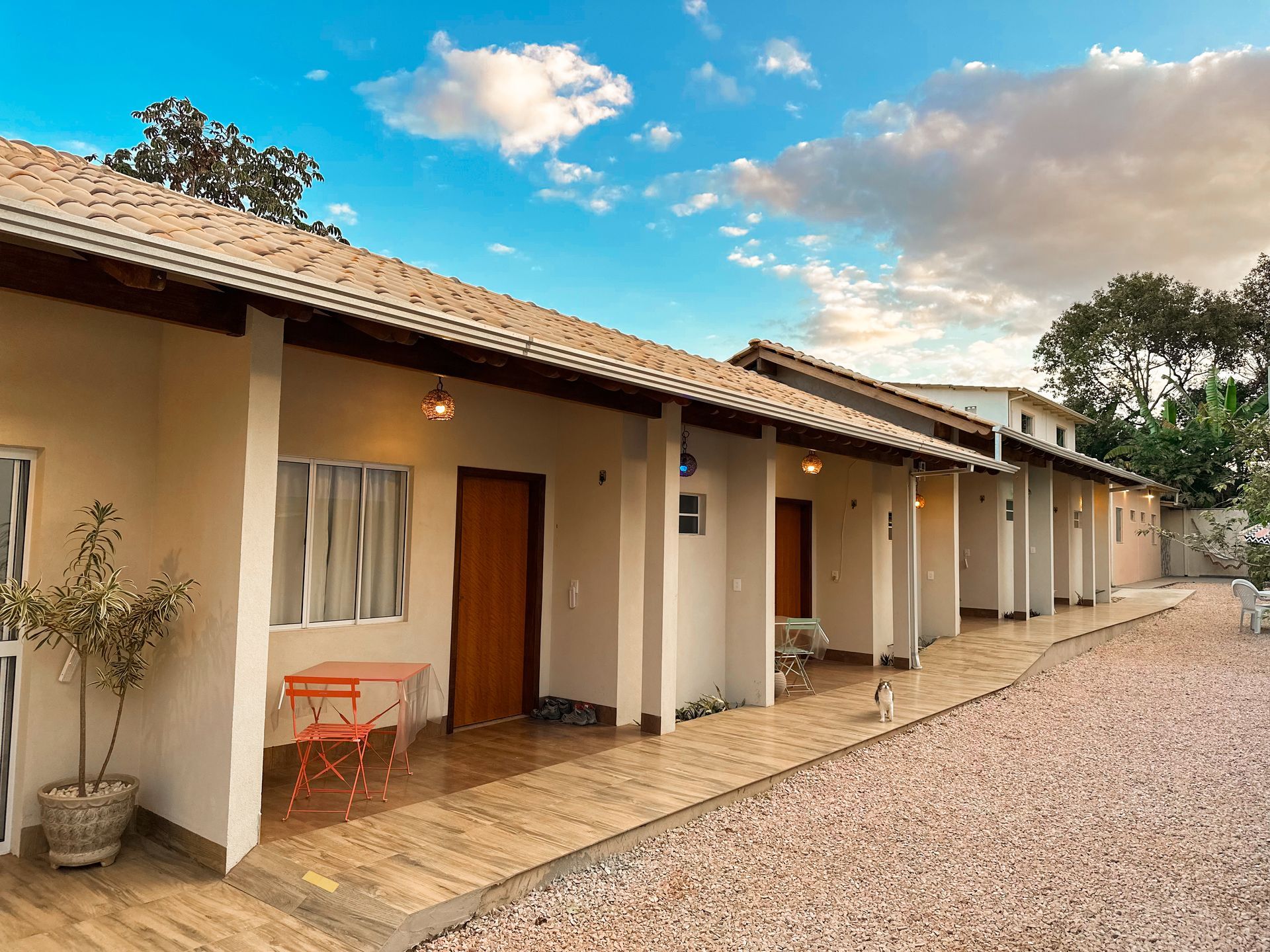 Fileira de quartos de hotel cor de bege com portas marrons, pequenas varandas e um caminho de cascalho sob um céu azul.