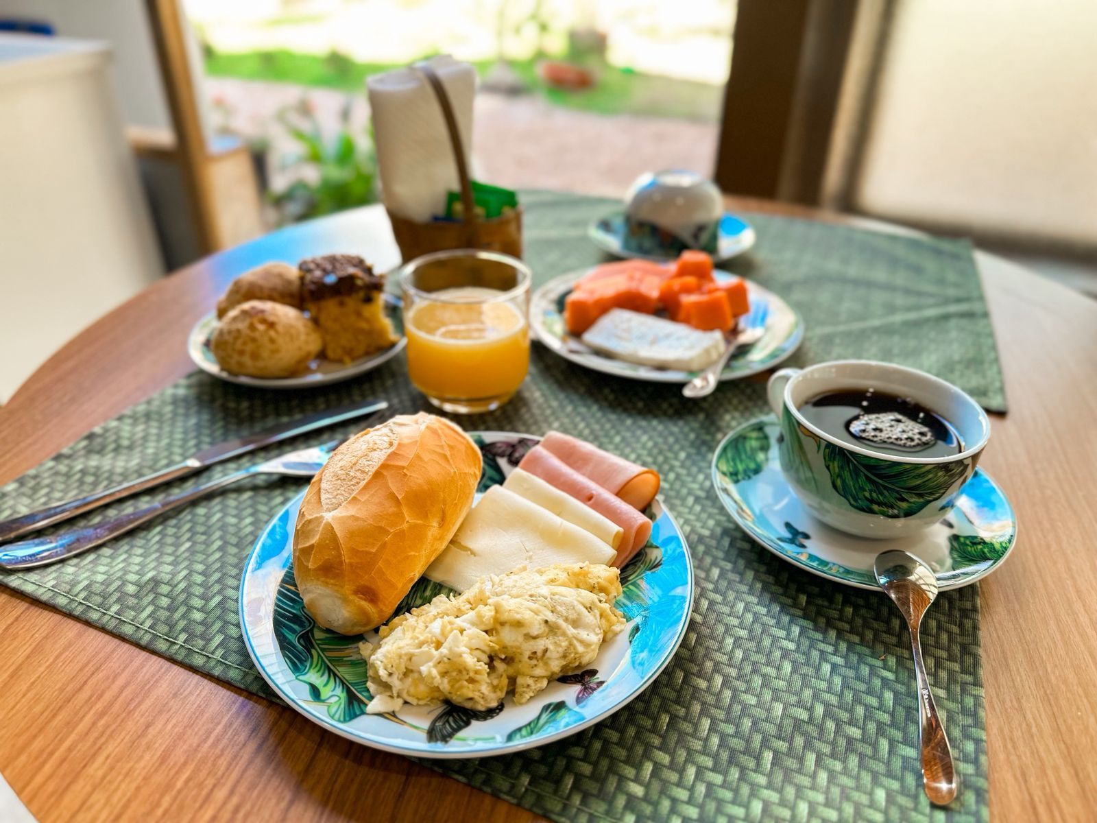 Café da manhã servido em uma mesa redonda de madeira: pão, café, suco, doces, frutas.