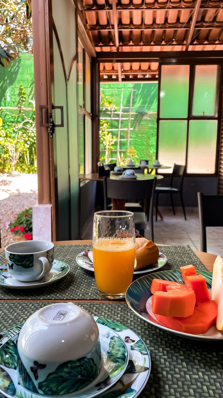 Mesa de café da manhã com suco, frutas e xícaras de café, com vista para um pátio ensolarado com mesa e cadeiras.