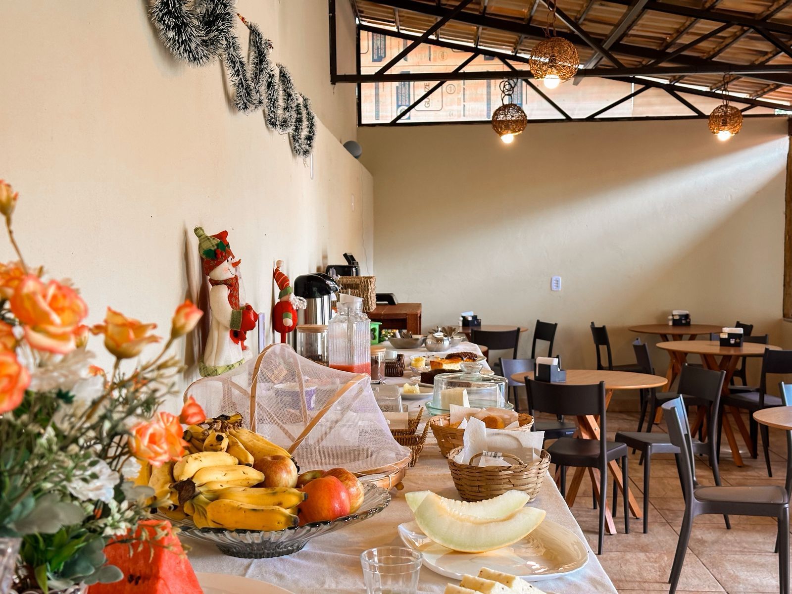 Buffet de café da manhã com frutas, doces e bebidas em uma mesa comprida; área de jantar com mesas e cadeiras.