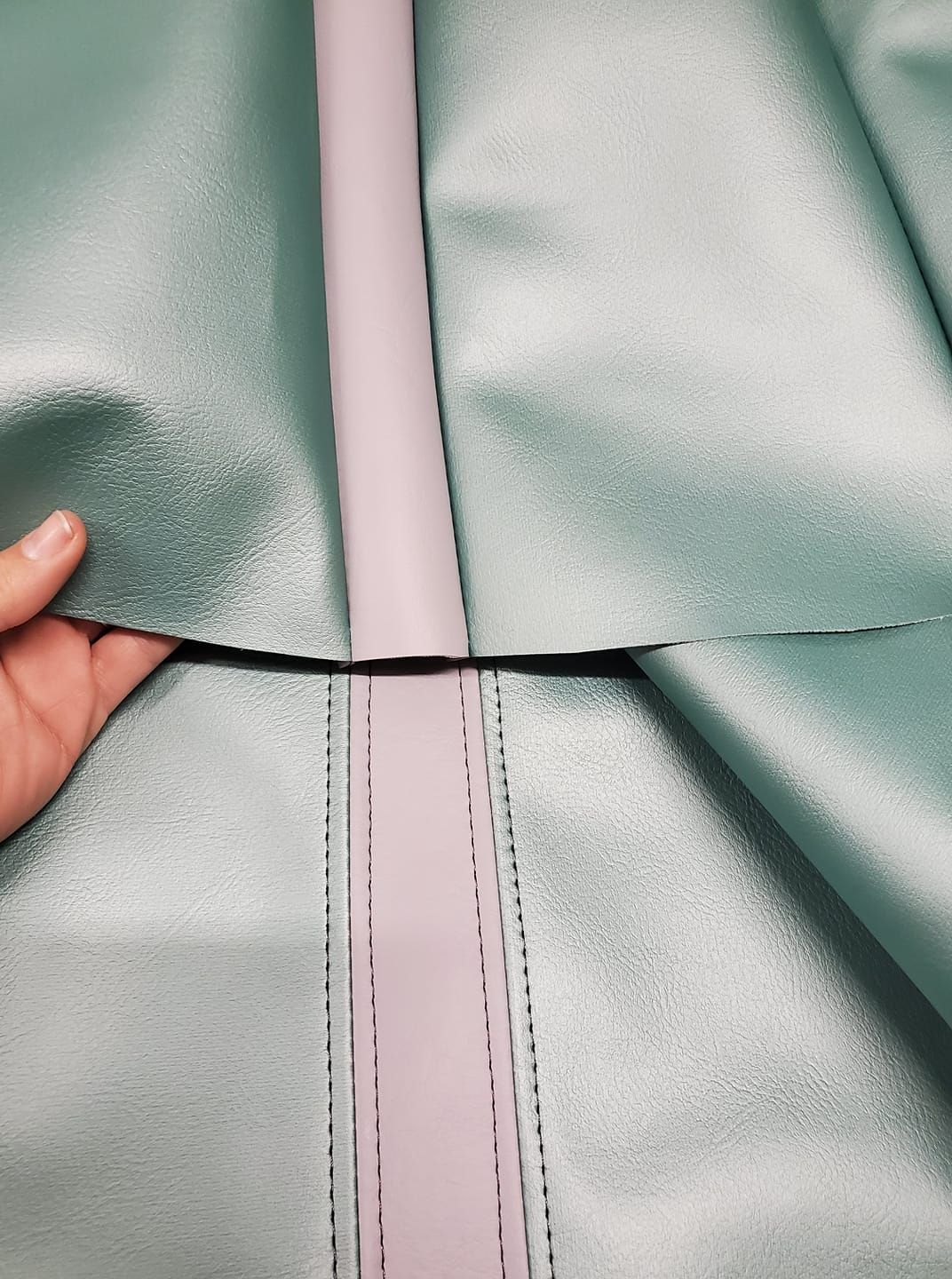 A hand holding up the edge of teal fabric, revealing a purple strip with stitching; indoors.