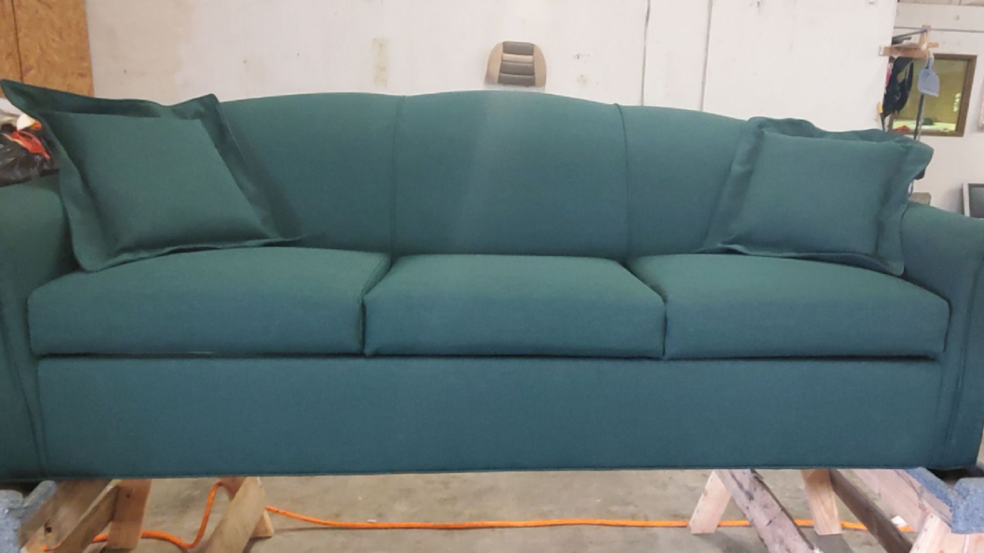 Teal sofa with two matching pillows. It is displayed in a workshop with a wooden frame underneath.