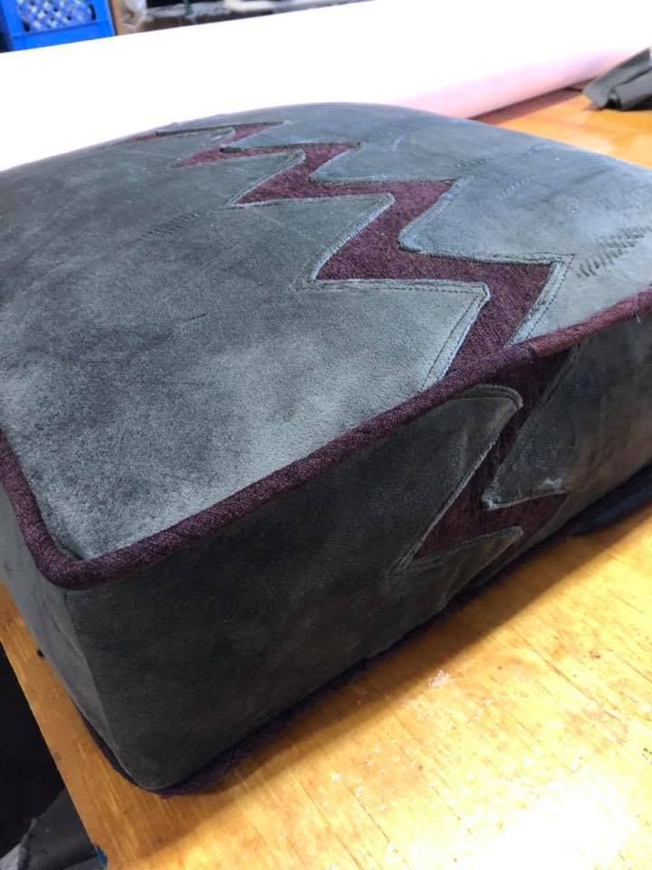 A square cushion with a green suede-like texture, maroon zig-zag design, and maroon trim sits on a wooden surface.