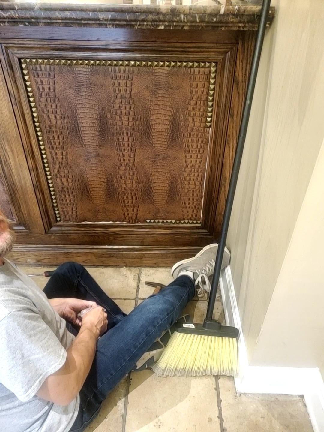 Person in jeans sitting against cabinet, broom leaning against wall.