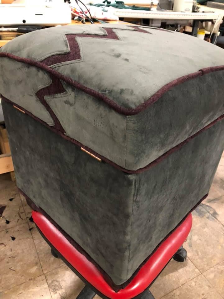 Square upholstered ottoman in gray-green fabric with burgundy trim and a zig-zag design, resting on a red chair.